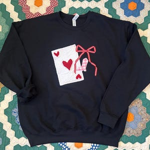 May include: Black sweatshirt featuring a playing card design with a red heart and a bow. The card is white with red hearts. The bow is red and white gingham. The sweatshirt is a crew neck style.