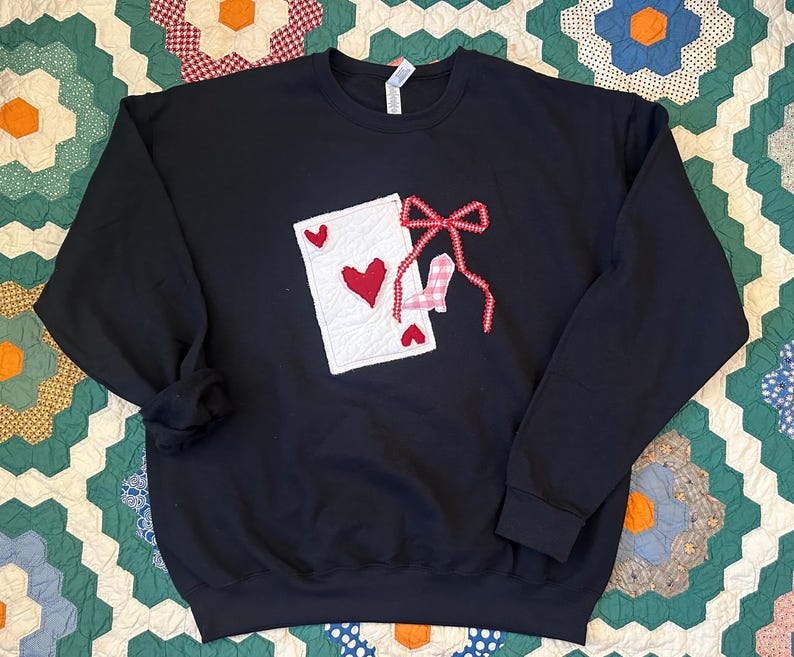 May include: Black sweatshirt featuring a playing card design with a red heart and a bow. The card is white with red hearts. The bow is red and white gingham. The sweatshirt is a crew neck style.
