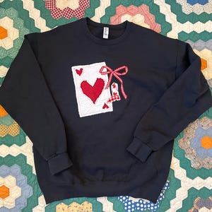 May include: A black sweatshirt with a playing card design featuring a red heart and a bow. The design is on the front of the sweatshirt. The sweatshirt is made of a soft material.