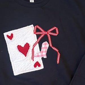 May include: Black sweatshirt with a decorative design featuring a playing card with red hearts, a red bow, and a pink and white gingham boot. The card is white with red hearts. The bow and boot are red and pink.