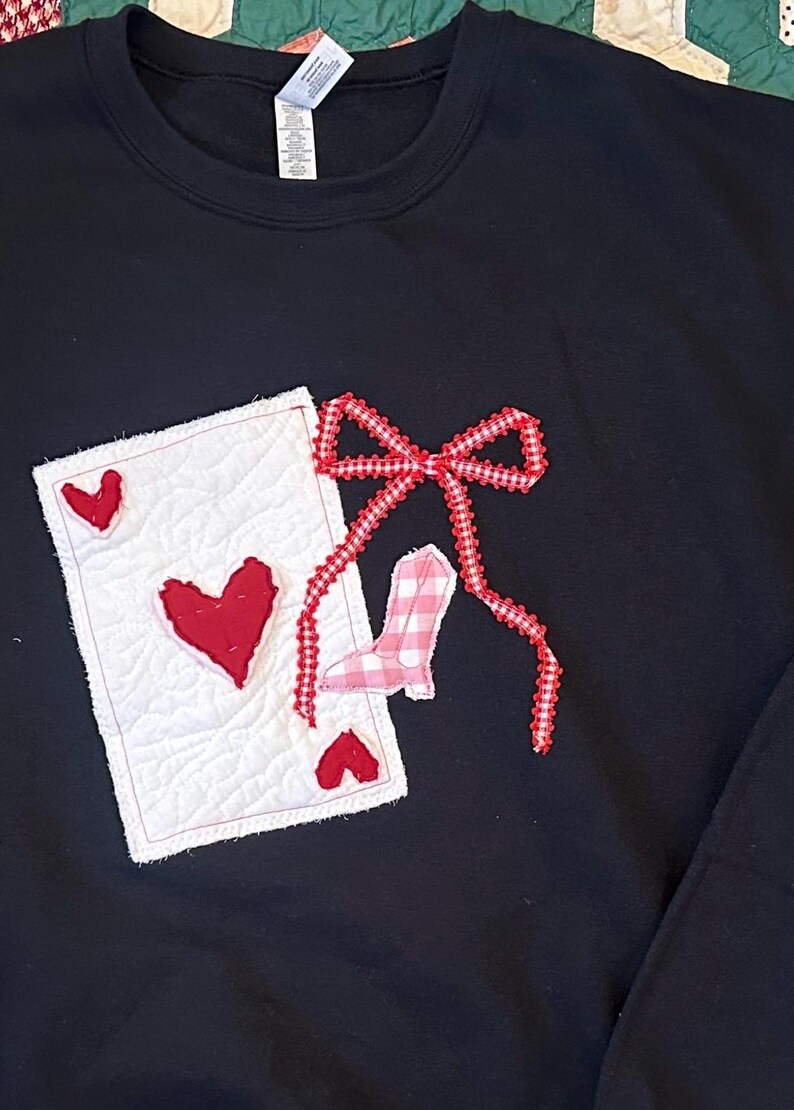 May include: Black sweatshirt with a decorative design featuring a playing card with red hearts, a red bow, and a pink and white gingham boot. The card is white with red hearts. The bow and boot are red and pink.
