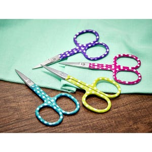 May include: Four pairs of small scissors with white polka dots on a green fabric background. The scissors are purple, pink, yellow, and blue.