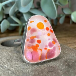May include: A silver ring with a pink, orange, and white glass stone. The stone has a speckled pattern.