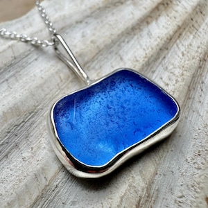 Könnte beinhalten: Eine silberne Halskette mit Anhänger, die einen rechteckigen, leuchtend blauen Seeglas-Anhänger zeigt. Das Seeglas ist in eine polierte Silberfassung gefasst und hängt an einer filigranen Silberkette. Der Anhänger hat eine glatte, strukturierte Oberfläche.
