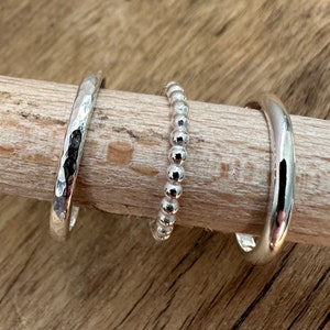 May include: Three silver rings on a wooden surface. The rings are different styles: one is hammered, one is beaded, and one is smooth.