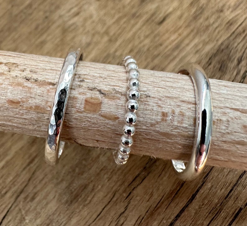 May include: Three silver rings on a wooden surface. The rings are different styles: one is hammered, one is beaded, and one is smooth.