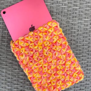 Könnte beinhalten: Ein pinkfarbenes Tablet, das teilweise in einer rechteckigen Häkelhülle steckt. Die Hülle weist ein strukturiertes Sternstichmuster aus mehrfarbigem Garn in leuchtendem Pink, Orange und Gelb auf, das den unteren Teil des Geräts bedeckt.