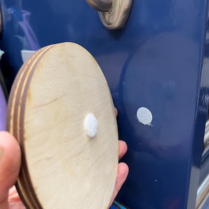 May include: A round wooden plaque with a white adhesive dot on the back. The plaque is made of multiple layers of wood and has a natural wood finish.