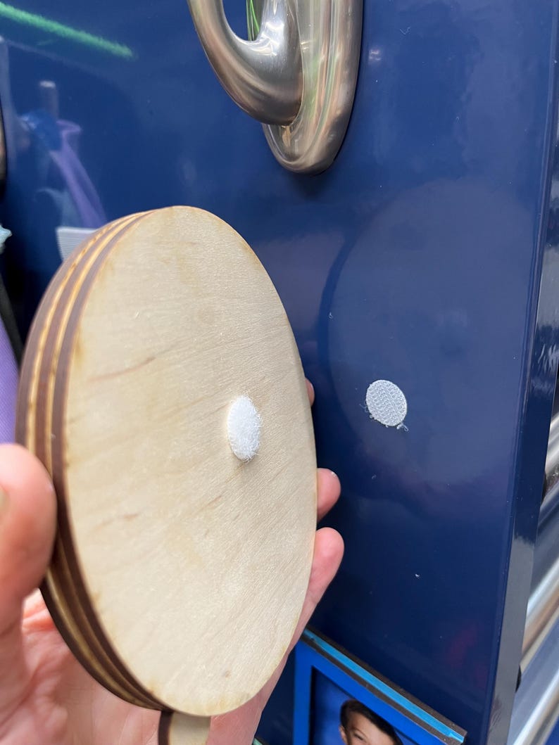 May include: A round wooden plaque with a white adhesive dot on the back. The plaque is made of multiple layers of wood and has a natural wood finish.