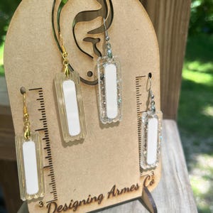 May include: Earrings displayed on a wooden earring holder. The earrings are rectangular with a white center and clear or glittery borders. The holder has a cutout ear shape and the text "Designing Armes Co."