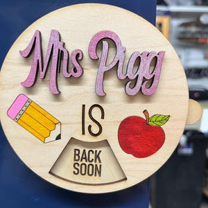 May include: A wooden sign with the text "Mrs. Pigg is back soon" in pink and black lettering. The sign also features a yellow pencil, a red apple, and a wooden sign that says "Back Soon".
