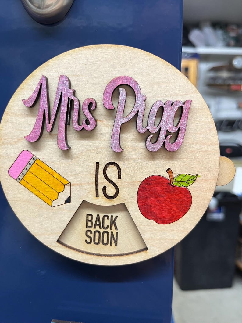 May include: A wooden sign with the text "Mrs. Pigg is back soon" in pink and black lettering. The sign also features a yellow pencil, a red apple, and a wooden sign that says "Back Soon".