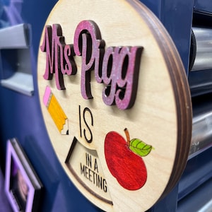 May include: A wooden sign with the text "Mrs. Pigg is in a meeting" in pink and black lettering. The sign also features a red apple and a yellow pencil.