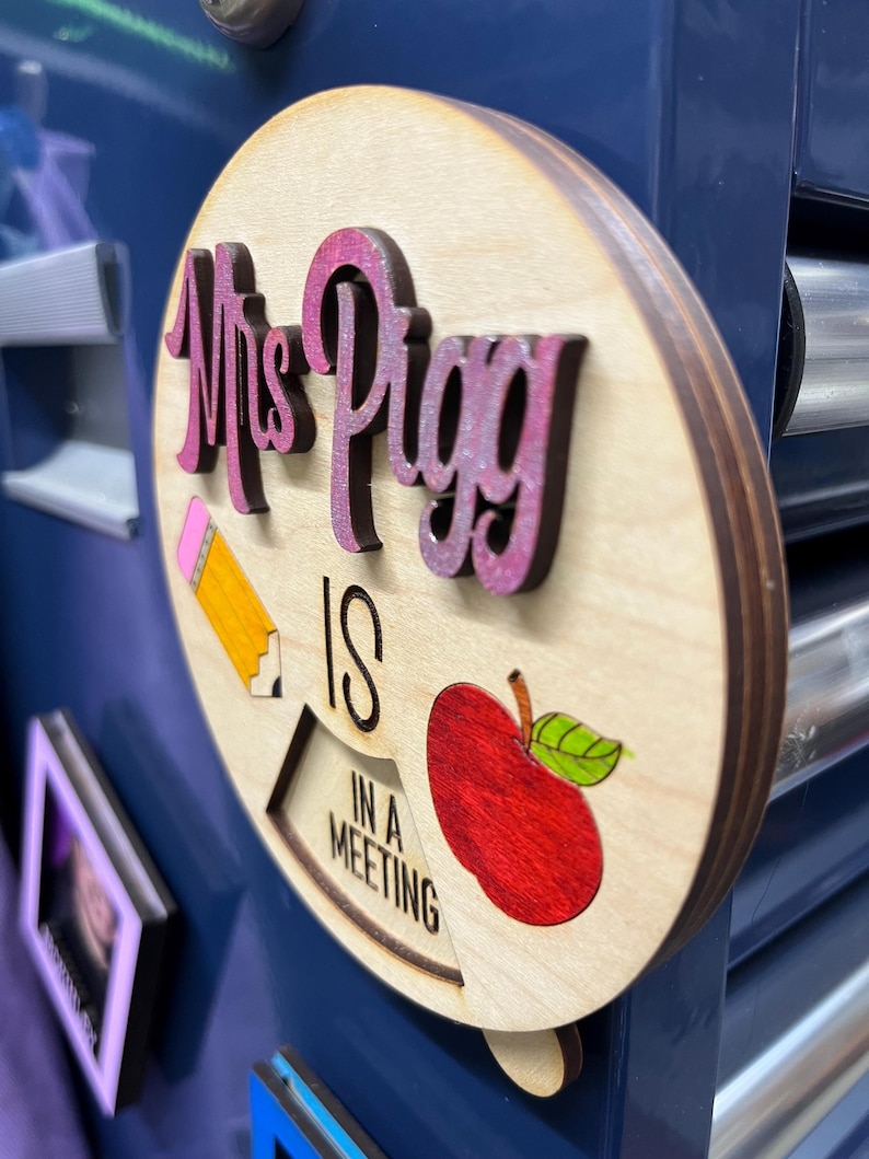 May include: A wooden sign with the text "Mrs. Pigg is in a meeting" in pink and black lettering. The sign also features a red apple and a yellow pencil.