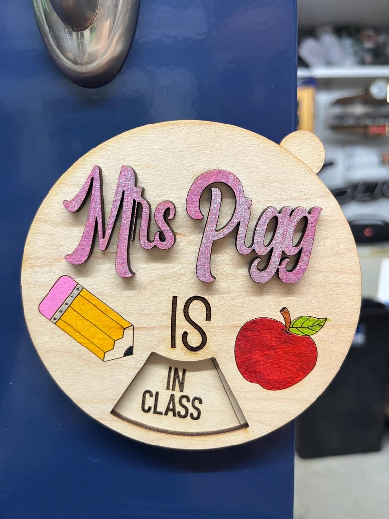 May include: A wooden sign with the text "Mrs. Pigg is in class" in pink and black lettering. The sign also features a red apple, a yellow pencil, and a rotating dial with the words "in class".