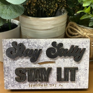 May include: Wooden sign with a white background and black lettering. The sign says "Stay Salty Stay Lit" with the scripture reference "Matthew 5:13-14" underneath.