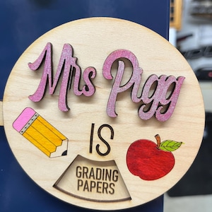 May include: A round wooden sign with the text "Mrs. Piggy is grading papers". The sign is decorated with a pink pencil, a red apple, and green leaves.