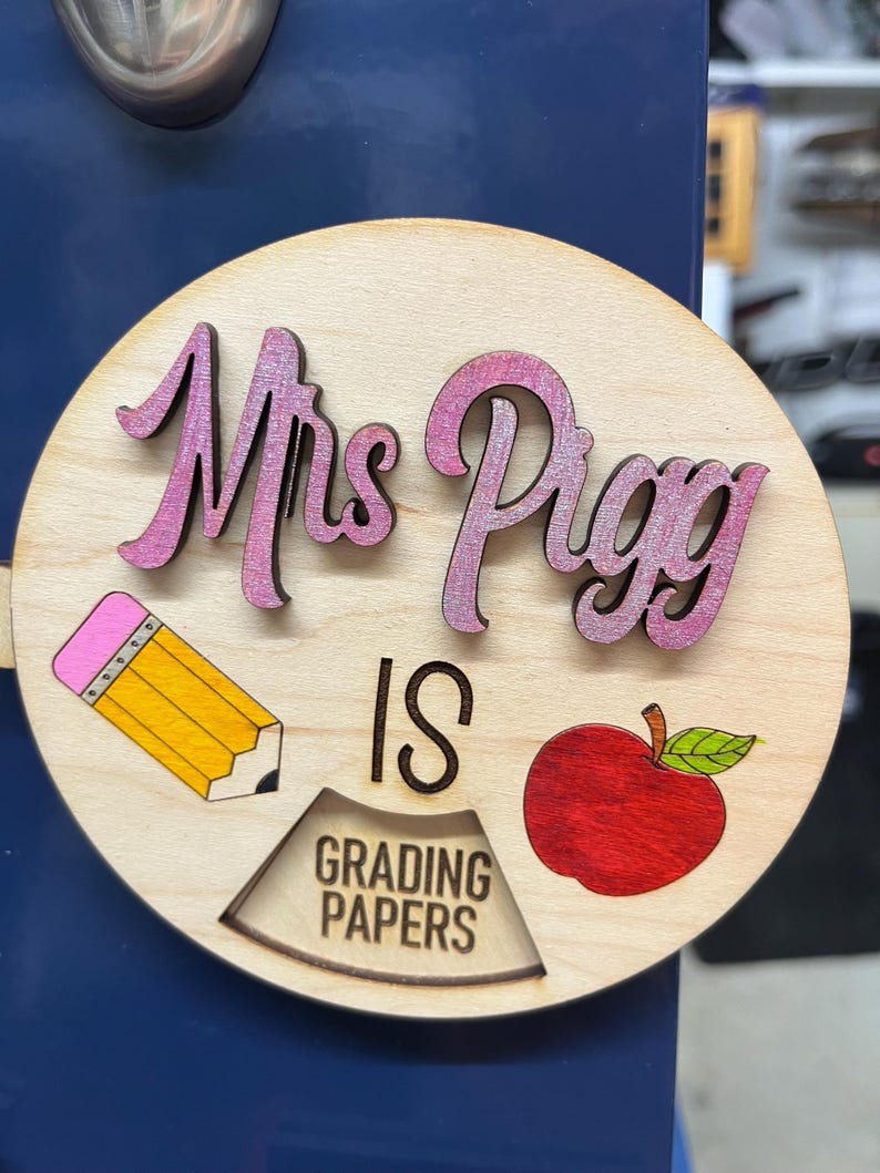 May include: A round wooden sign with the text "Mrs. Piggy is grading papers". The sign is decorated with a pink pencil, a red apple, and green leaves.