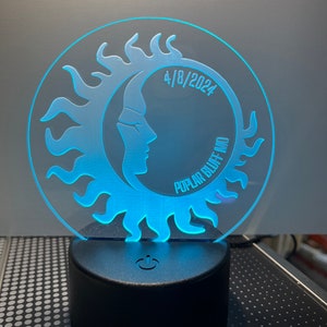 May include: A circular, illuminated night light featuring a sun and moon design with the text "4/8/2024" and "Poplar Bluff MO". The light emits a soft blue glow and sits on a black base with a power symbol.