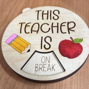 May include: A wooden sign with the text "This Teacher Is On Break" in black lettering. The sign is decorated with a yellow pencil and a red apple.