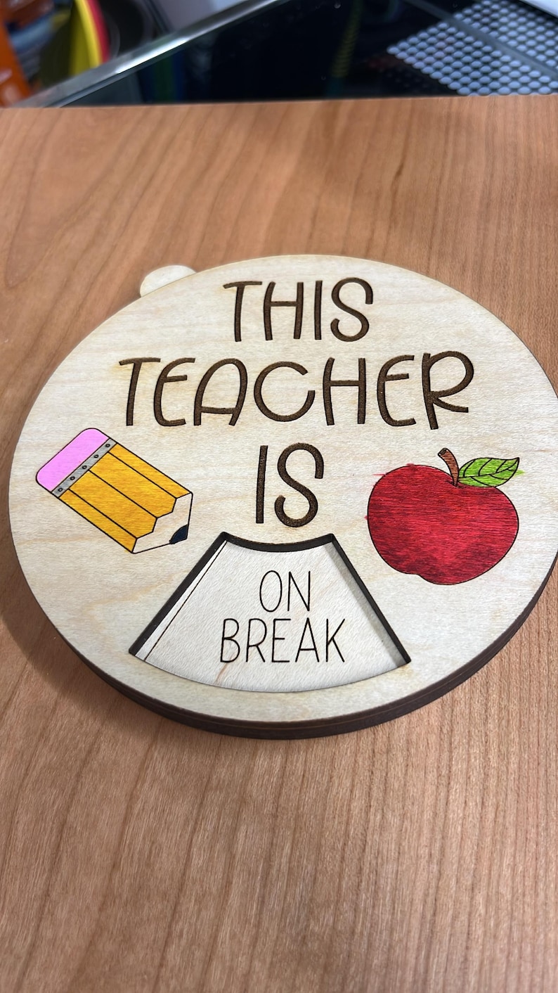 May include: A wooden sign with the text "This Teacher Is On Break" in black lettering. The sign is decorated with a yellow pencil and a red apple.