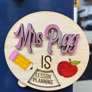 May include: A round wooden sign with the text "Mrs. Piggy is Lesson Planning" in pink and black lettering. The sign also features a red apple, a yellow pencil, and a pink eraser.