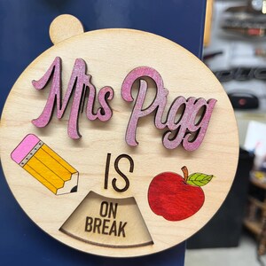 May include: A round wooden sign with the text "Mrs. Pigg is on break" in black lettering. The sign also features a pink pencil, a red apple, and a pink and white "Mrs. Pigg" in cursive.