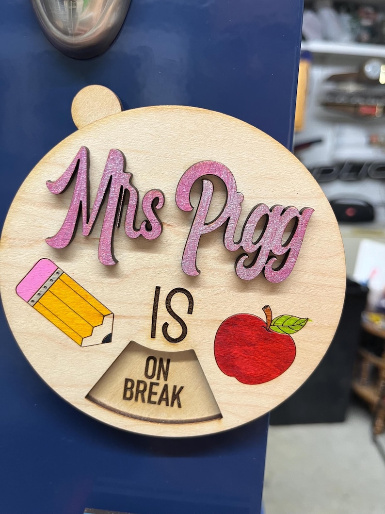 May include: A round wooden sign with the text "Mrs. Pigg is on break" in black lettering. The sign also features a pink pencil, a red apple, and a pink and white "Mrs. Pigg" in cursive.