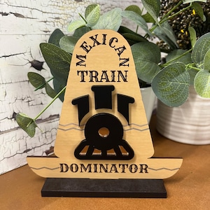 May include: Wooden sign with the words "MEXICAN TRAIN DOMINATOR" and a black train graphic. The sign is light brown with black text and a black base. The sign is a decorative item.
