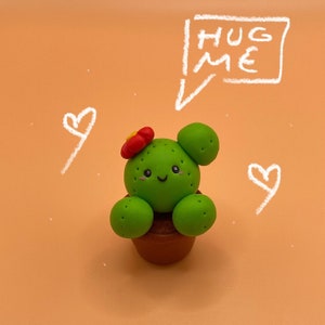 Könnte beinhalten: Ein grüner und roter Tonkaktus mit einem lächelnden Gesicht und einer roten Blüte oben. Der Kaktus steht in einem braunen Topf und hat die Worte "Hug Me" in Weiß darüber geschrieben.