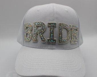 May include: A light gray baseball cap with a white brim. The word "BRIDE" is spelled out in large, iridescent rhinestone letters across the front of the cap. The cap is a classic six-panel design.