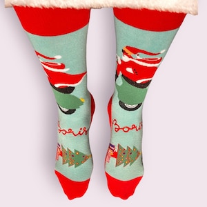 May include: A pair of Christmas-themed socks with a red cuff and toe. The socks feature a light blue background with Santa Claus riding a green scooter and a Christmas tree design. The word "Boris" is printed on the socks.