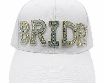 May include: A white baseball cap featuring the word BRIDE in large, block-style letters across the front. Each letter is filled with numerous small, iridescent, multi-colored rhinestones that reflect light, creating a shimmering effect against the fabric.