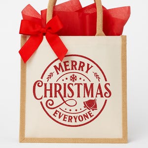 May include: A beige tote bag with a red "Merry Christmas Everyone" design. The bag has a jute trim and handles, a red bow, and red tissue paper inside. The design is a circular shape with a bell and snowflake.