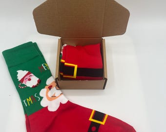 May include: Christmas-themed socks, one green and one red, with Santa Claus designs. The red sock features a black belt and buckle. The socks are displayed with a small cardboard box, one pair folded inside and another pair lying outside.