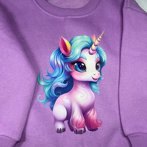 May include: Purple sweatshirt featuring a colourful cartoon unicorn graphic. The unicorn has blue hair, pink and white fur, and a gold horn.