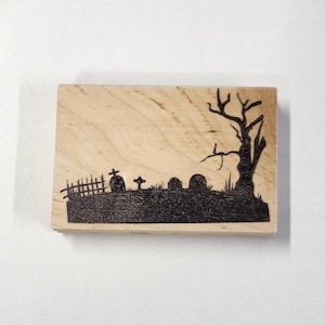 May include: A wooden stamp with a black silhouette of a graveyard with a fence, crosses, tombstones, and a bare tree.