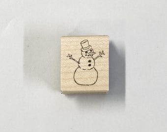 Pode incluir: Um carimbo de madeira com um desenho de linha de um boneco de neve usando um chapéu e com um nariz de cenoura.