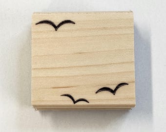 以下が含まれることがあります： 明るい木目模様の正方形の木製スタンプ。3つの黒い鳥のシルエットが表面にエッチングされており、2つは下部に、1つは上部にあります。スタンプはクラフトツールです。