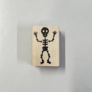 May include: A wooden stamp with a black silhouette of a skeleton with a skull head, arms raised, and feet together.
