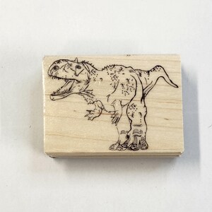 May include: A wooden rubber stamp with a black and white line drawing of a dinosaur. The dinosaur is standing on all fours with its mouth open.
