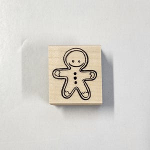 May include: A wooden stamp featuring a gingerbread man design. The gingerbread man is outlined in black, with a smiling face and three button details. The stamp is rectangular and made of light-colored wood.