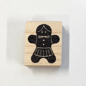 May include: A wooden block stamp with a black gingerbread person design. The gingerbread person is wearing a skirt and a bow.