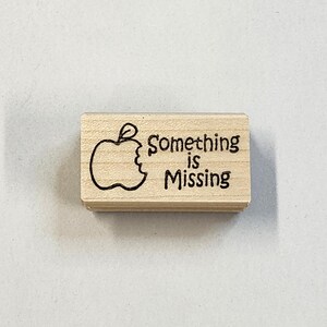 Op de afbeelding: Houten stempel met een zwarte omtrek van een gebeten appel en de tekst "Something is Missing".