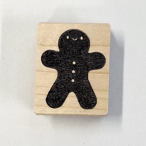 May include: A wooden stamp with a black gingerbread person design. The gingerbread person has a smiling face and three buttons.