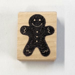 May include: A wooden stamp with a black gingerbread man design. The gingerbread man is smiling and has buttons on its shirt.