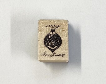 以下が含まれることがあります： クリスマスのオーナメントのデザインが施された木製のスタンプ。スタンプには手書きのフォントで「merry christmas」と書いてあります。