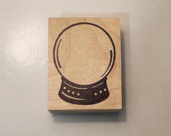 Pode incluir: Um carimbo de madeira com uma ilustração preta de uma bola de cristal. A bola de cristal é mostrada com um globo de vidro circular e uma base decorativa. O carimbo é feito de madeira clara.