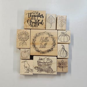 May include: A set of 11 wooden stamps with various autumn-themed designs, including a sunflower, a pumpkin, a gourd, a basket of apples, a leaf, and a wreath with the text "Hello Fall".