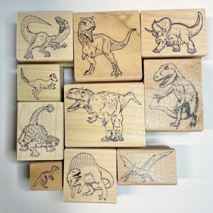 Op de afbeelding: Een set van negen houten dinosaurus stempels. De stempels tonen verschillende dinosaurussen, waaronder een T-Rex, een Triceratops en een Stegosaurus. De stempels zijn allemaal in zwart-wit en perfect voor knutselen en scrapbooking.
